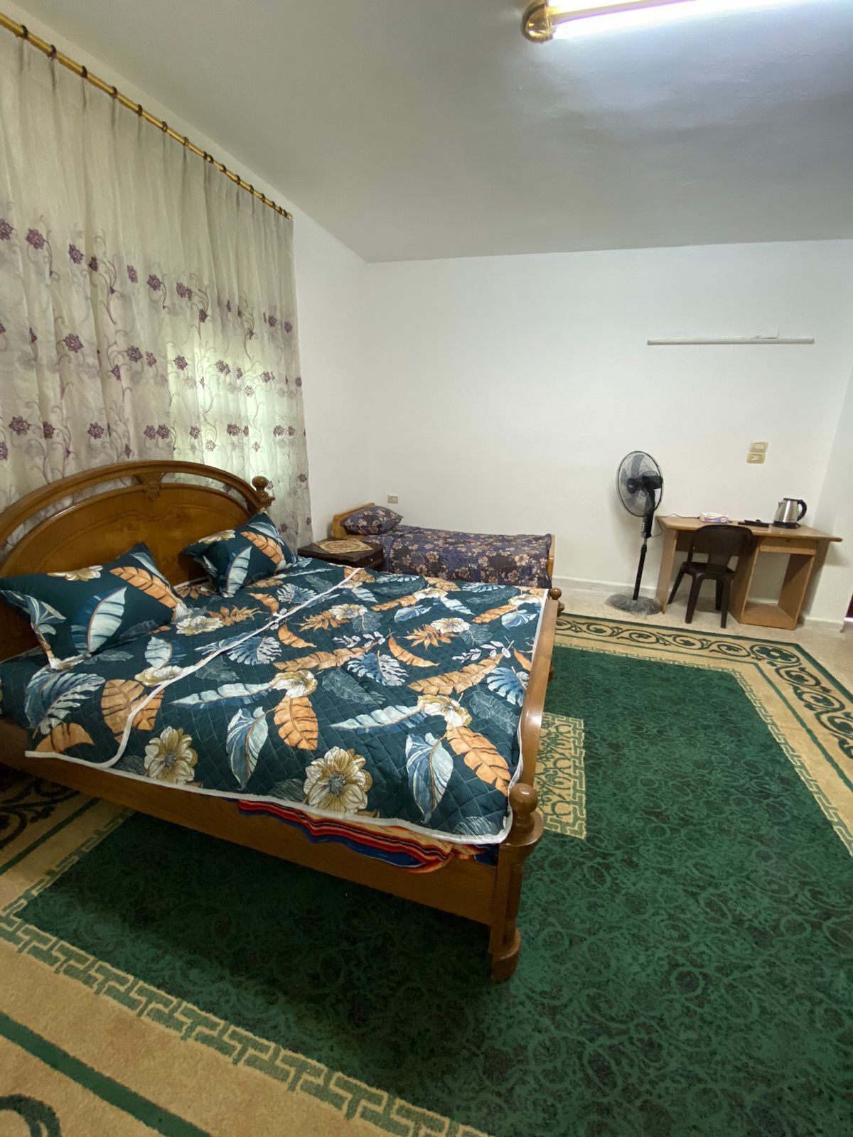 Une chambre à coucher avec un lit, un bureau, une chaise, un ventilateur et un rideau.