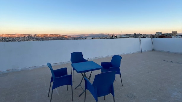 A rooftop terrace with blue chairs and a table, overlooking a cityscape during sunset.