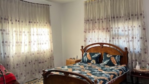 A bedroom with a wooden bed, floral curtains, a green patterned rug, and a ceiling light.