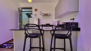 A modern kitchen with two bar stools, a refrigerator, a coffee maker, and a microwave. - Cozy Studio just 15 minutes to Karura Forest & 5 Minutes to Two Rivers Mall. (Ruaka)