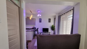 A living room with a dark sofa, a TV, and a small kitchen area with a microwave and bar stools. - Cozy Studio just 15 minutes to Karura Forest & 5 Minutes to Two Rivers Mall. (Ruaka)