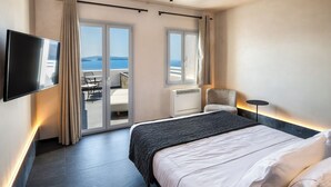 Quarto de hotel moderno com uma cama grande, televisão de tela plana, uma cadeira e vista para o mar pelas portas deslizantes.