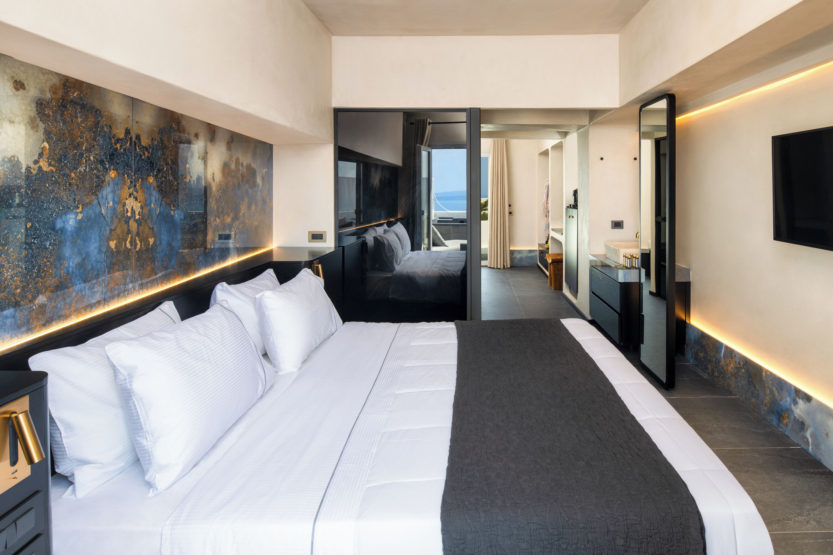 A modern hotel room with a large bed, a view of the sea, a bathroom with a mirror, and a large abstract painting on the wall.