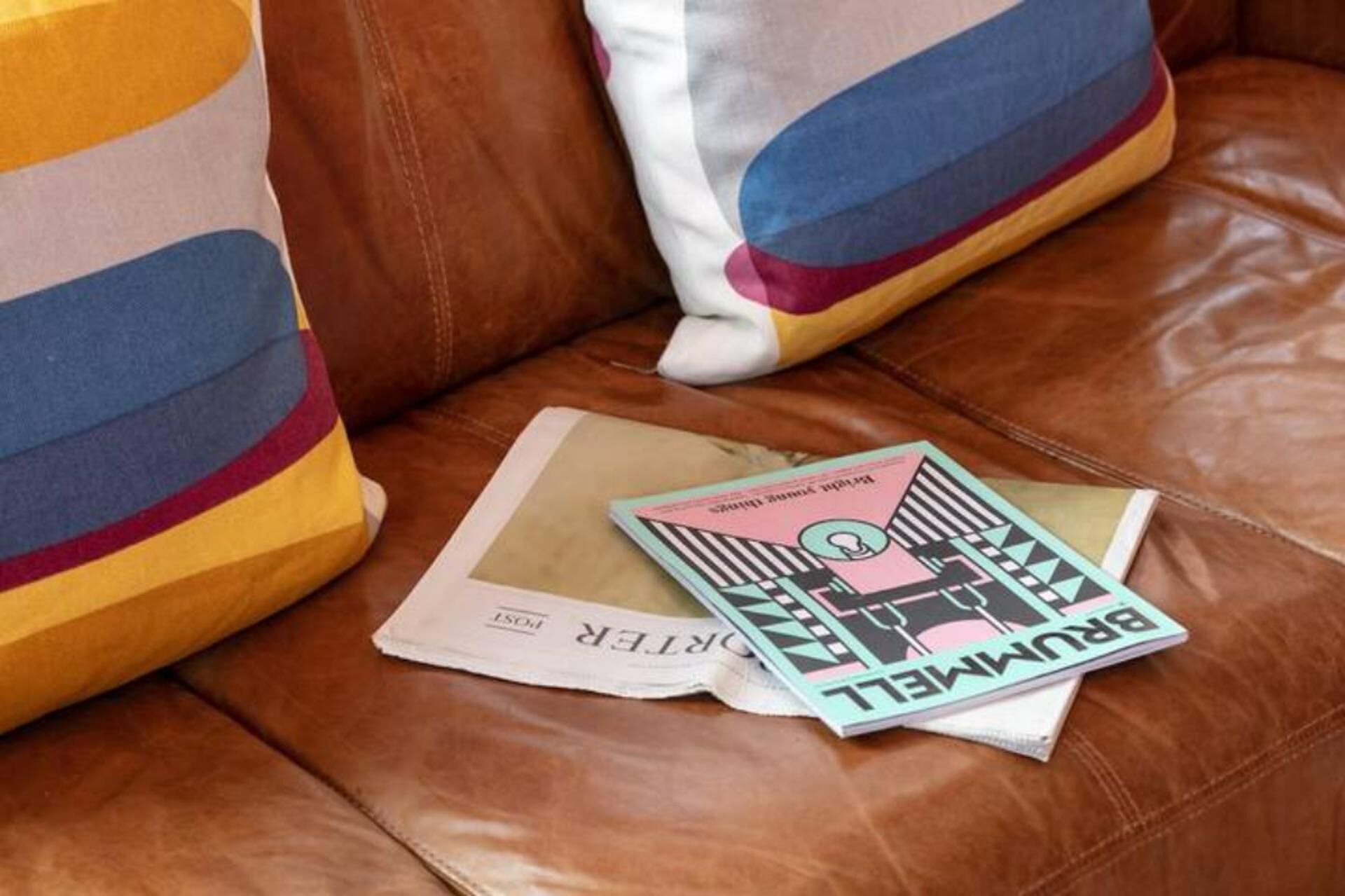 A brown leather sofa with colorful pillows and a magazine on it.