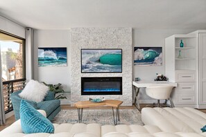 A modern living room with a fireplace, a television mounted on a textured wall, and a view of the ocean through large windows. - Fairway Haven - Luxury Carlsbad Golf Course Condo on the Omni La Costa Resort (Carlsbad)