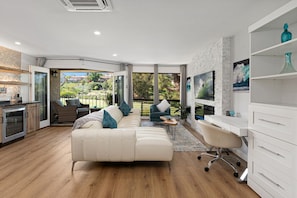 A modern living room with a sofa, chairs, a desk, and a television. There is a kitchen area with a stove and cabinets. The room has large windows and a view of greenery outside. - Fairway Haven - Luxury Carlsbad Golf Course Condo on the Omni La Costa Resort (Carlsbad)