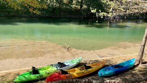 Una hilera de kayaks alineados en una orilla arenosa junto a una superficie acuática tranquila, con árboles al fondo.