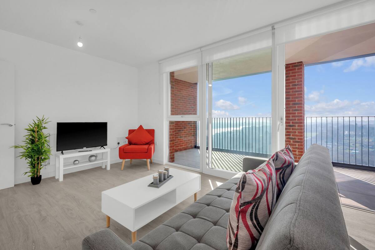 Un salon moderne avec un téléviseur à écran plat, une table basse blanche, un canapé gris et un fauteuil rouge. Il y a un balcon avec vue sur l’océan.