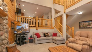 Una sala de estar acogedora con un sofá modular, una mesa de centro de madera y una pared de piedra. Un televisor está montado en la pared, y una escalera conduce a un nivel superior.