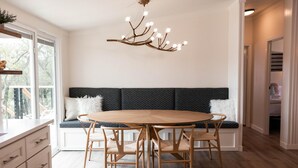 Uma área de refeições com uma mesa redonda de madeira, quatro cadeiras, um banco estofado em padrão preto e branco, e um lustre moderno.