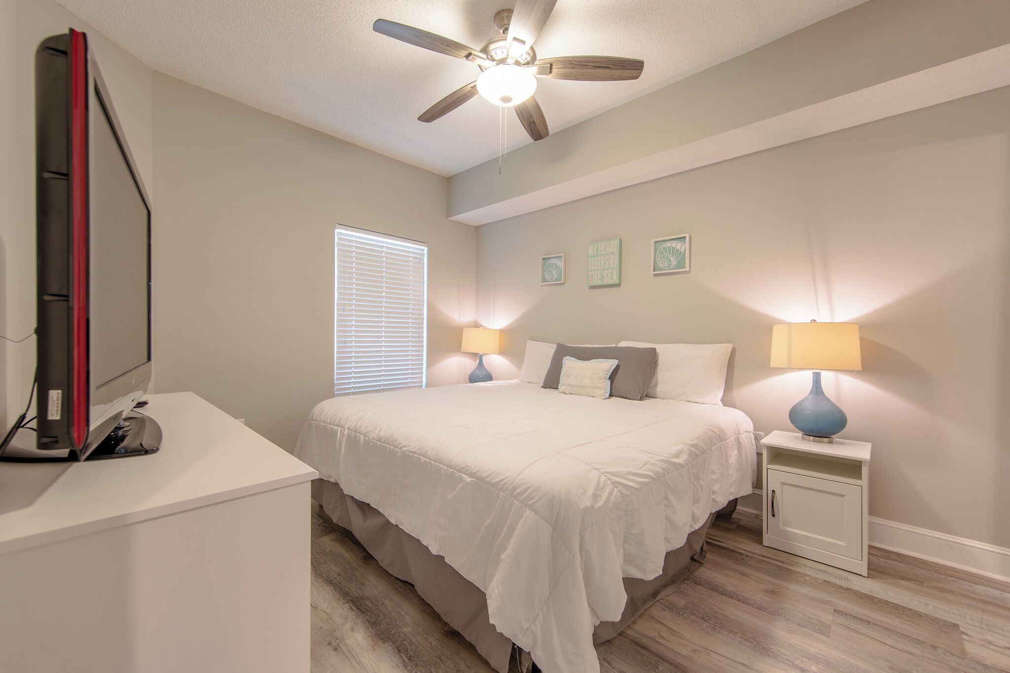 A bedroom with a bed, bedside lamps, a ceiling fan, a desk with a computer, and a window with blinds.