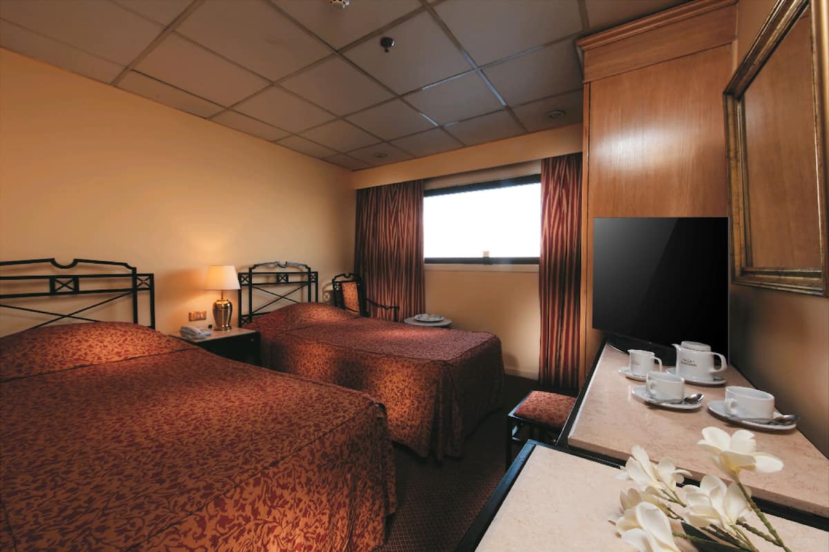 Quarto de hotel com duas camas, uma televisão, uma mesa com xícaras e pires, e uma janela com cortinas.