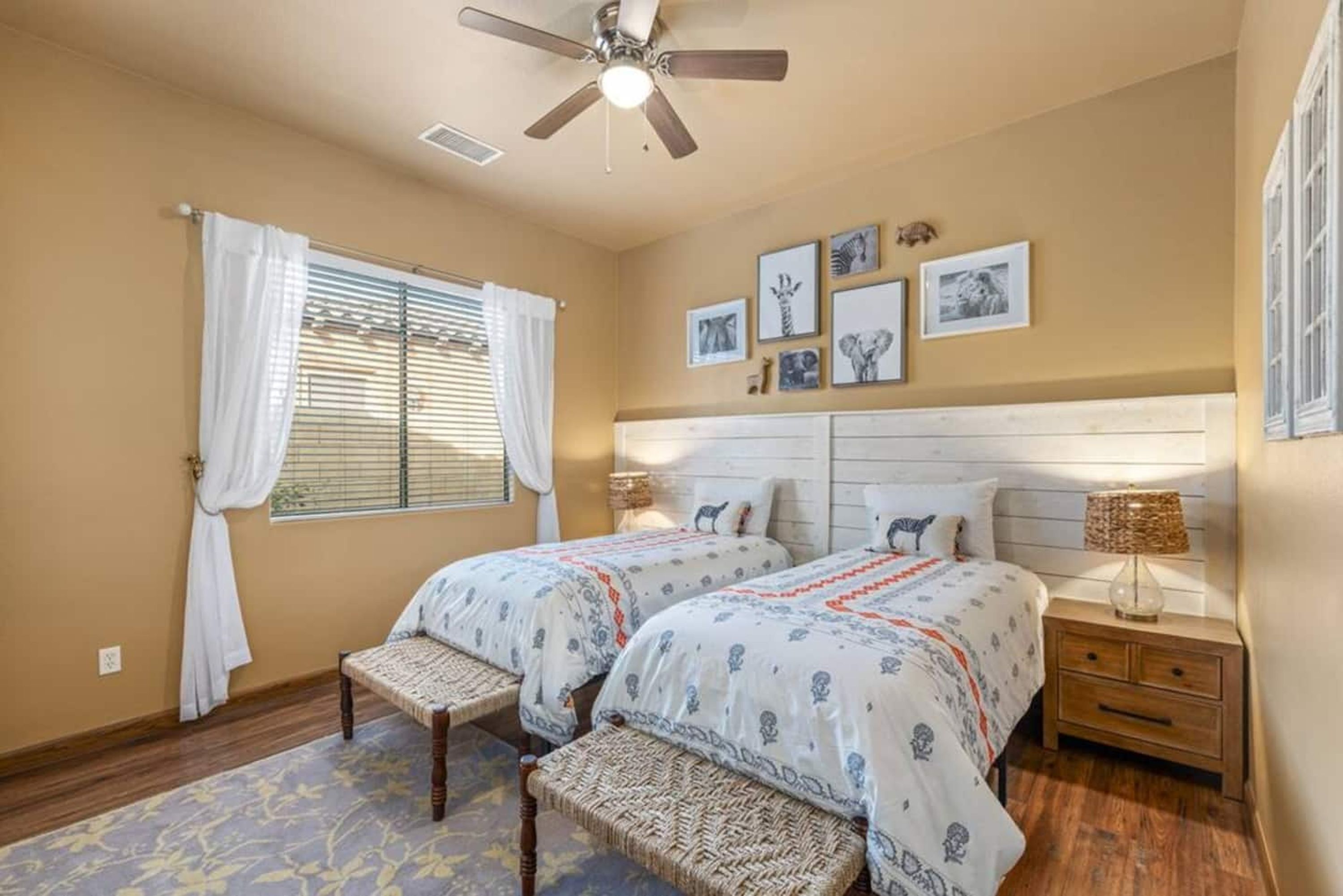 A bedroom with two beds, a ceiling fan, a window with curtains, and wall-mounted pictures.