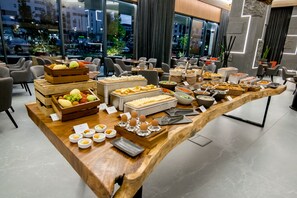 A buffet setup with various food items and utensils on a wooden table, set in a modern dining area with chairs and a view of the cityscape through large windows.