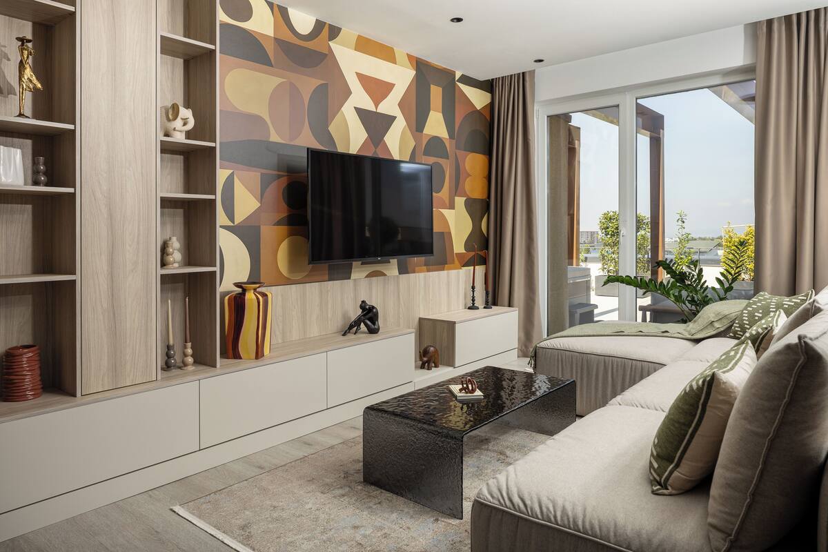 A modern living room with a flat-screen TV mounted on a geometric-patterned wall, a sofa, a coffee table, and a bookshelf with decorative items.