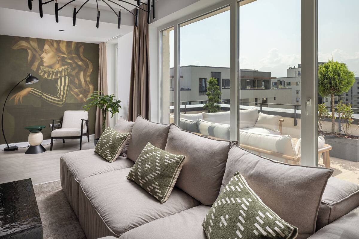 A modern living room with a beige sofa, green patterned pillows, a side table, a floor lamp, and a large window offering a view of the cityscape.