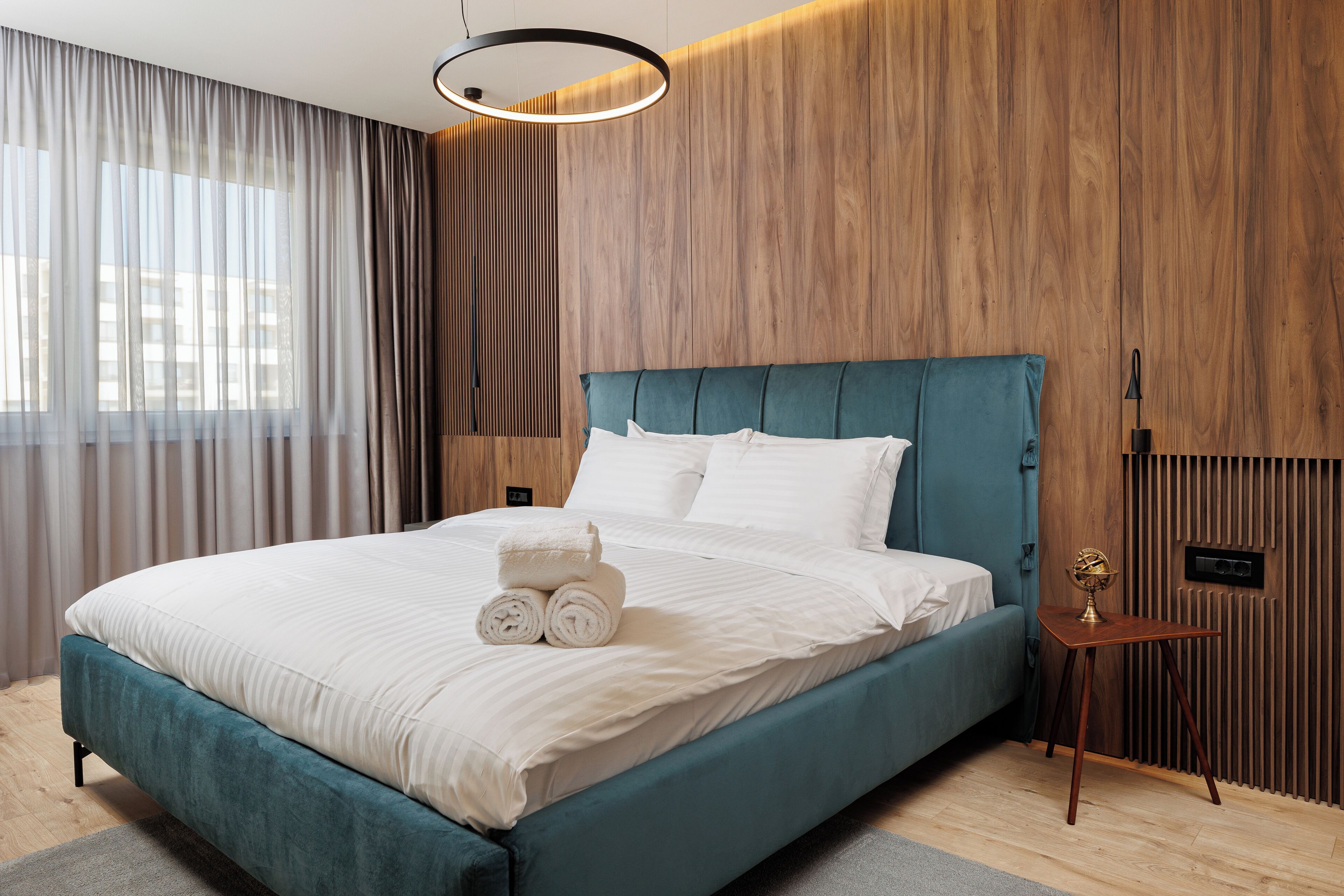 A modern hotel room with a teal bed, white linens, a wooden panel wall, and a small bedside table with a lamp.