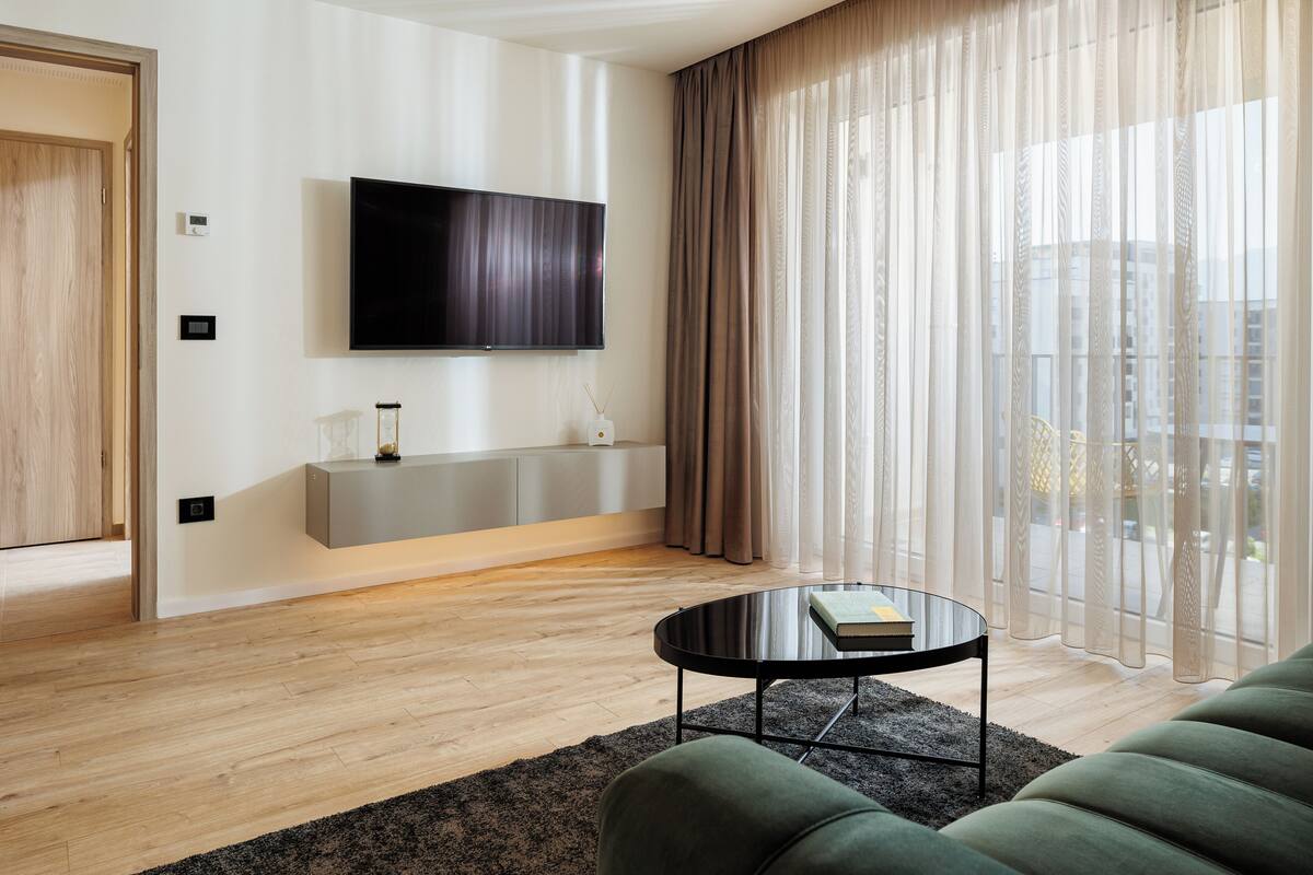 A modern living room with a flat-screen TV mounted on the wall, a grey console table, a round coffee table, and a green sofa.
