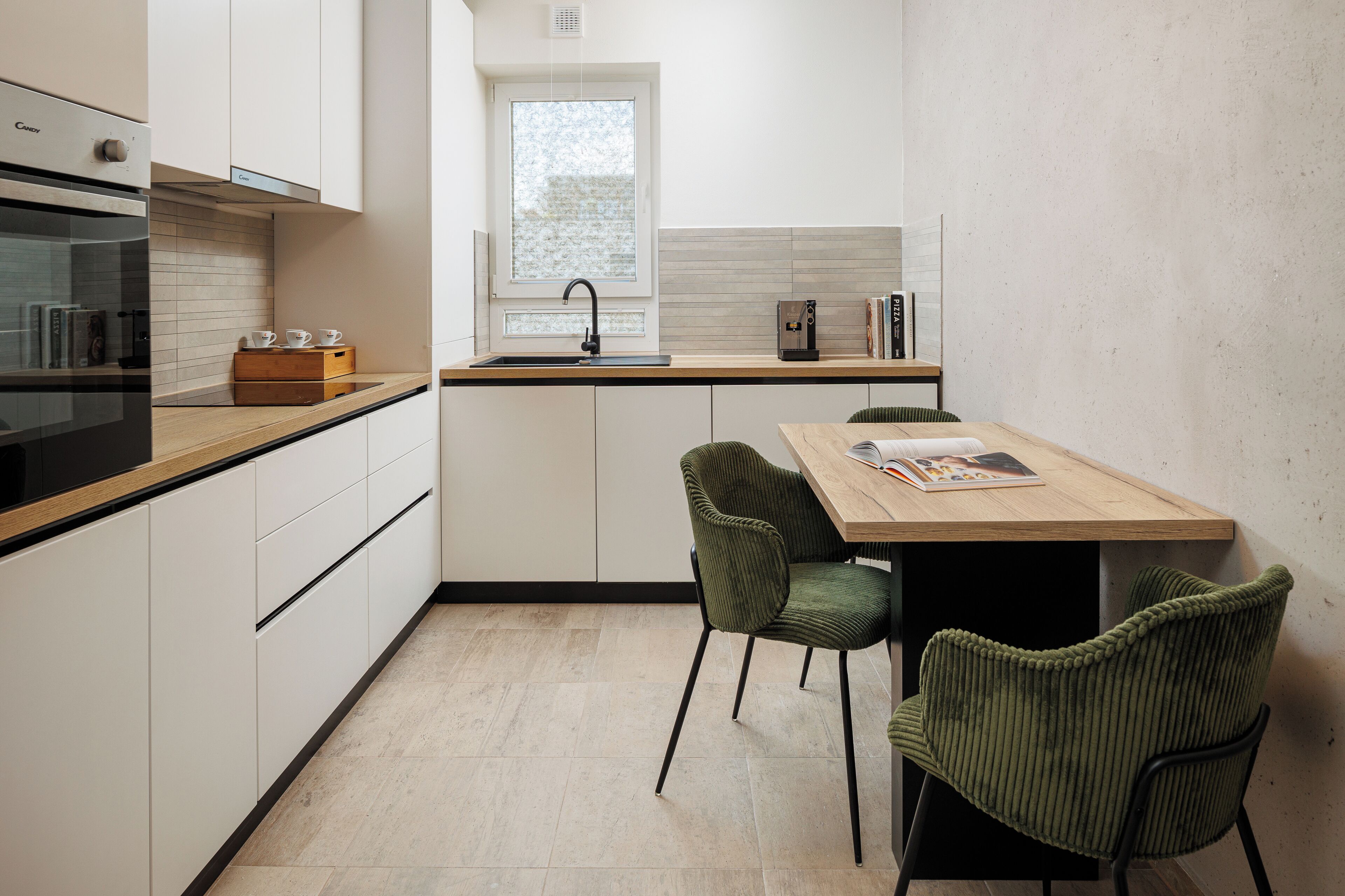 A modern kitchen with white cabinetry, a wooden countertop, a built-in oven, a window, and two green chairs.