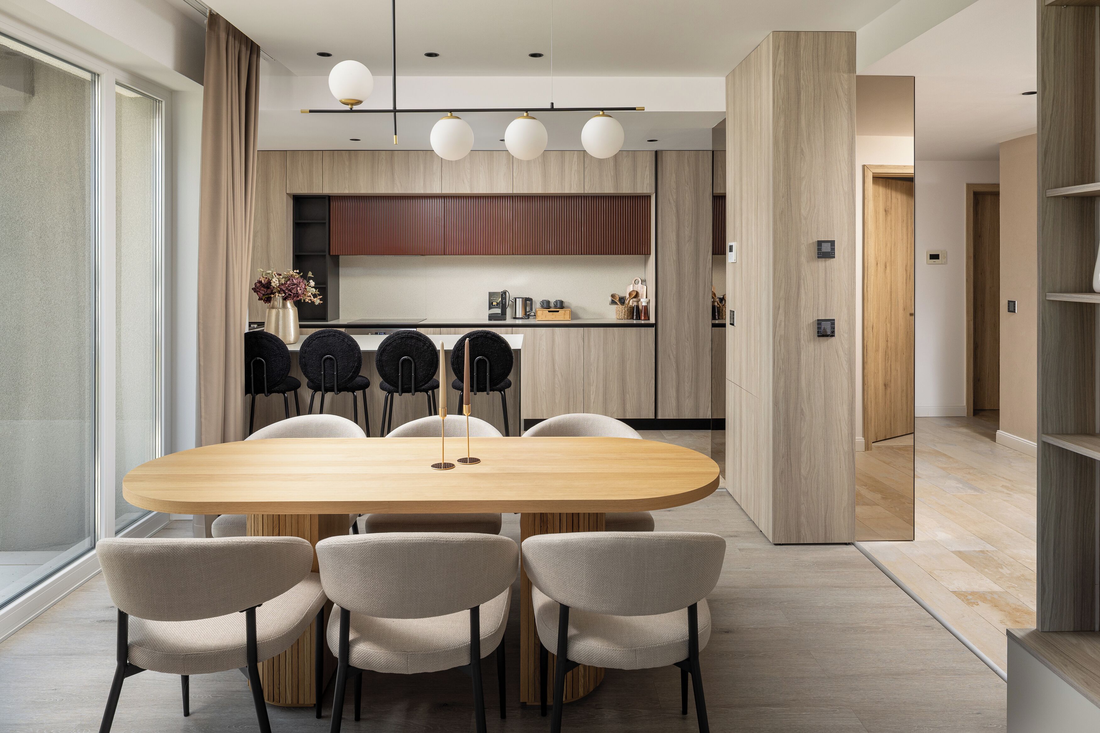 A modern dining area with a wooden table and four chairs, a kitchen in the background, and a large window on the left.