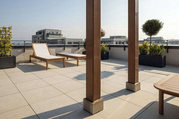 A rooftop terrace with sun loungers, potted plants, and a view of surrounding buildings.