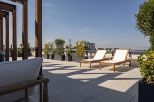 Terraza en la azotea con dos sillones de madera y una butaca acolchada, rodeada de macetas con plantas y una vista despejada del horizonte urbano.