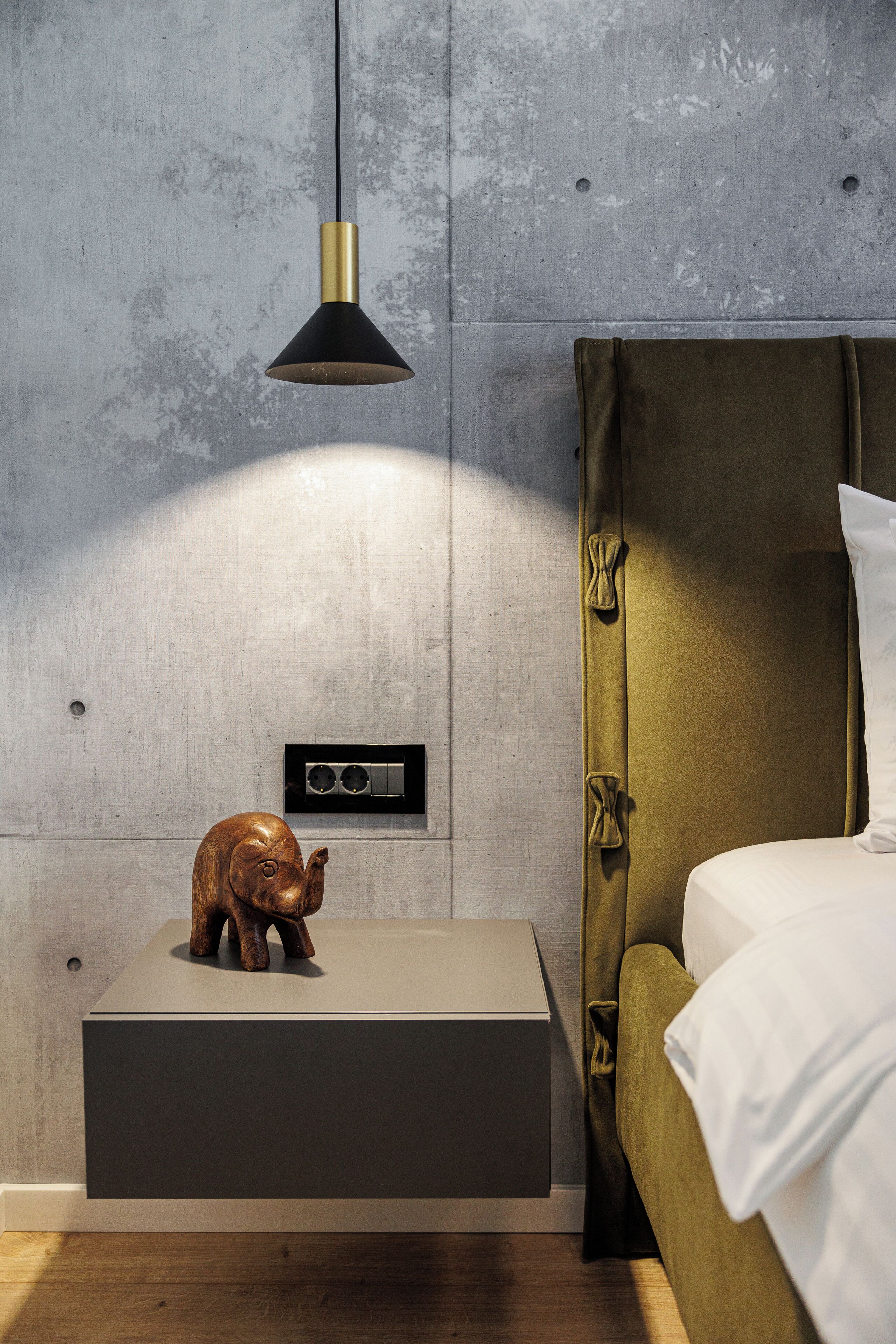 Camera da letto moderna dal design minimalista, con un letto dotato di una testiera verde, un comodino con una scultura di un elefante in legno e una lampada a sospensione nera.