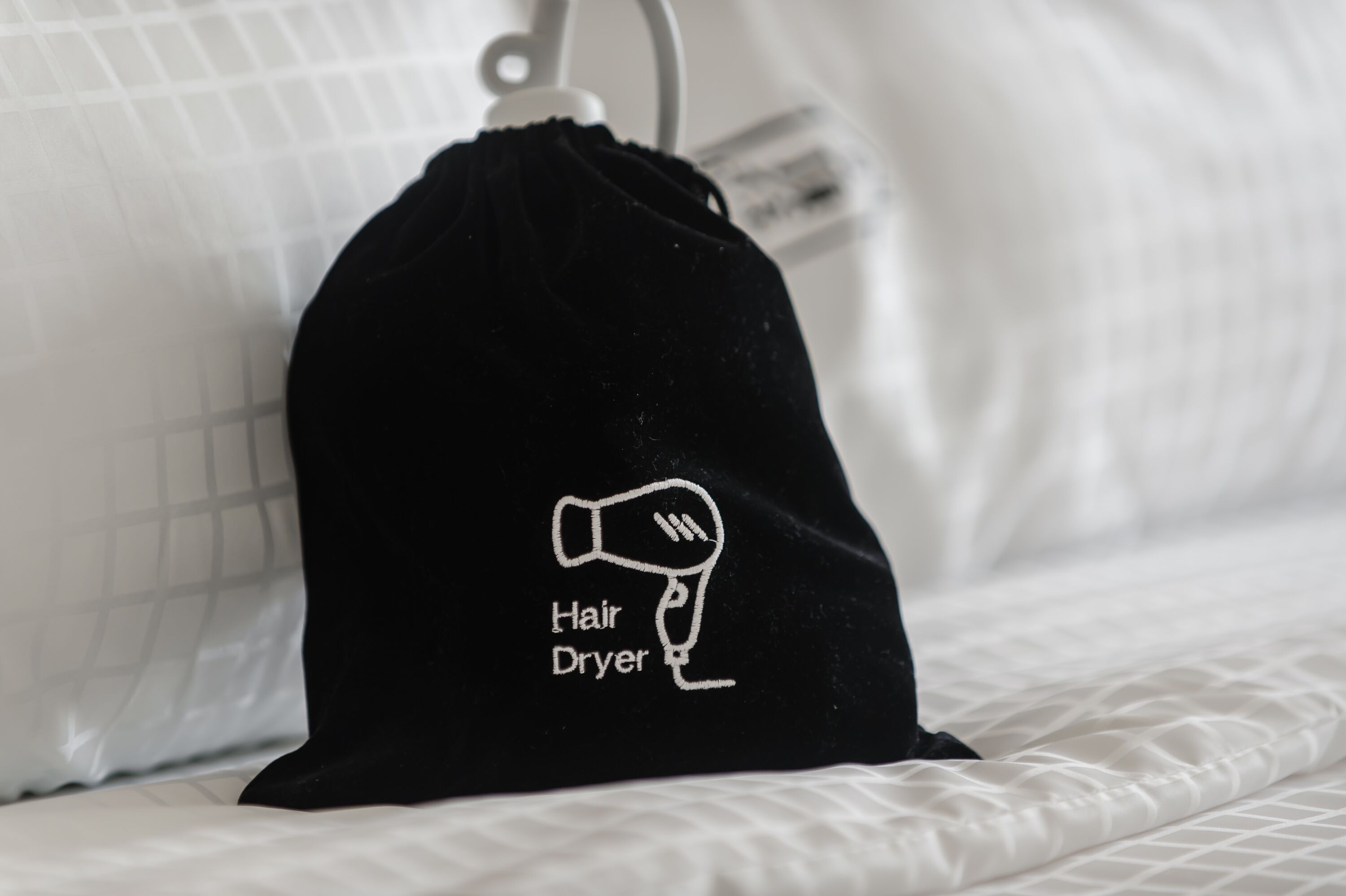 A black drawstring bag with a white hair dryer icon and the text "Hair Dryer" on a bed with white bedding.