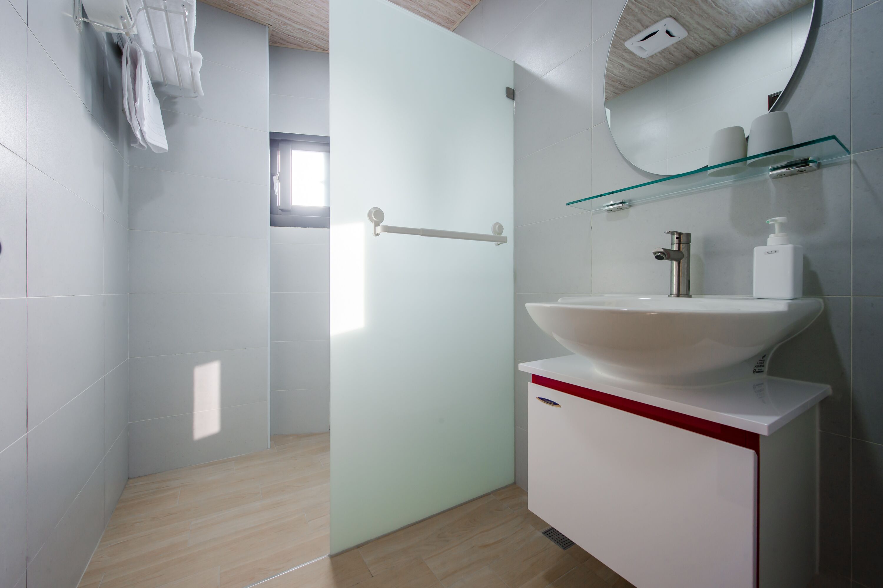 Un baño moderno con repisas de vidrio que sostienen artículos de aseo, un lavamanos blanco y un perchero.