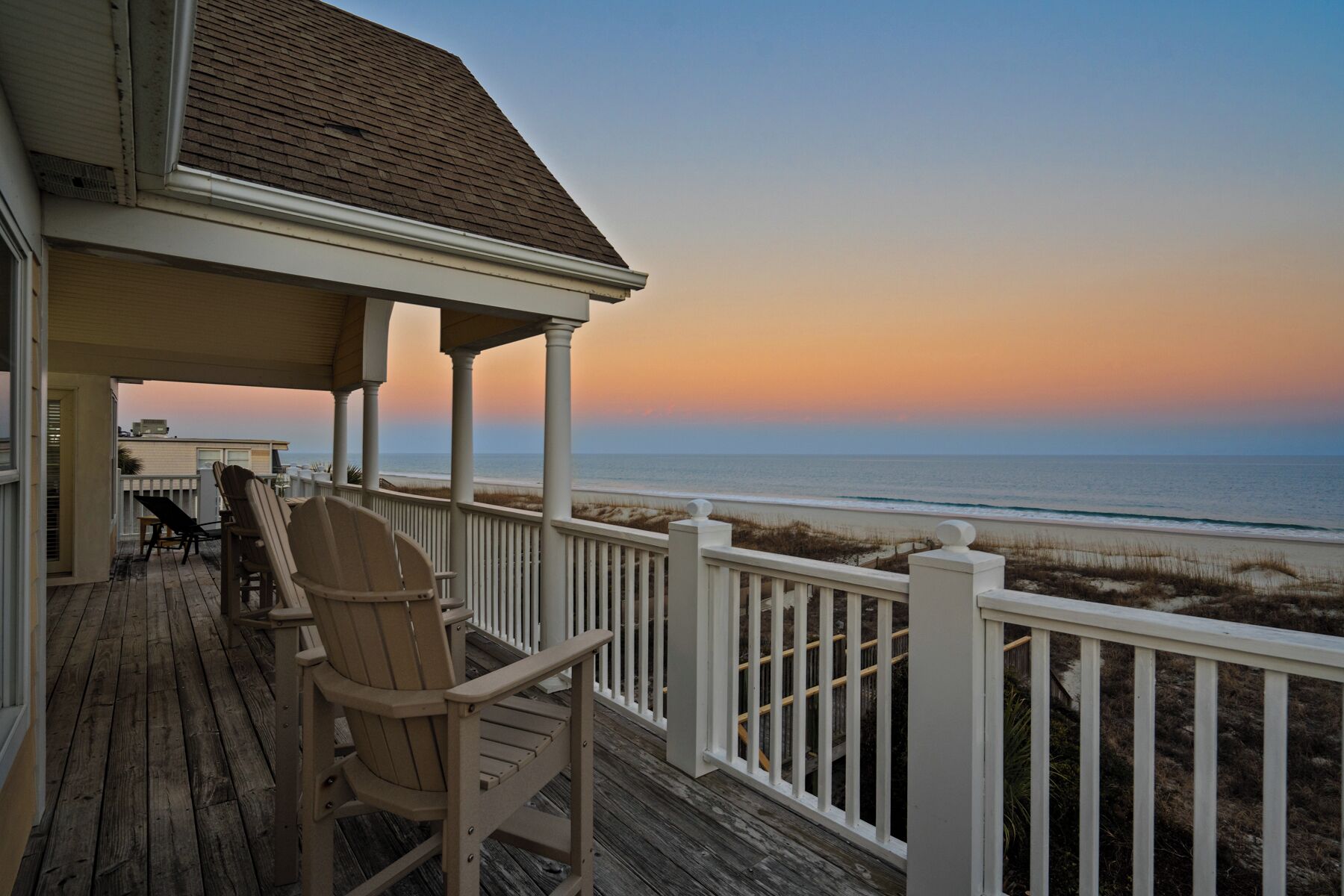 Een houten terras met stoelen en een tafel, met uitzicht op een strand bij zonsondergang.
