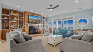 A living room with a fireplace, a television, and a view of the ocean through large windows. - Marlin View: Oceanfront w/ Pool, Walk to Dining (Garden City)
