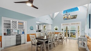 A spacious kitchen with white cabinets, a dining area with wooden chairs, and a view of the outdoors through large windows. - Marlin View: Oceanfront w/ Pool, Walk to Dining (Garden City)