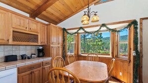 A wooden kitchen with a dining table and chairs, wooden cabinets, and a window with a view of trees. - Monterey Mtn View Cabin  Views Moonridge (Big Bear Lake)