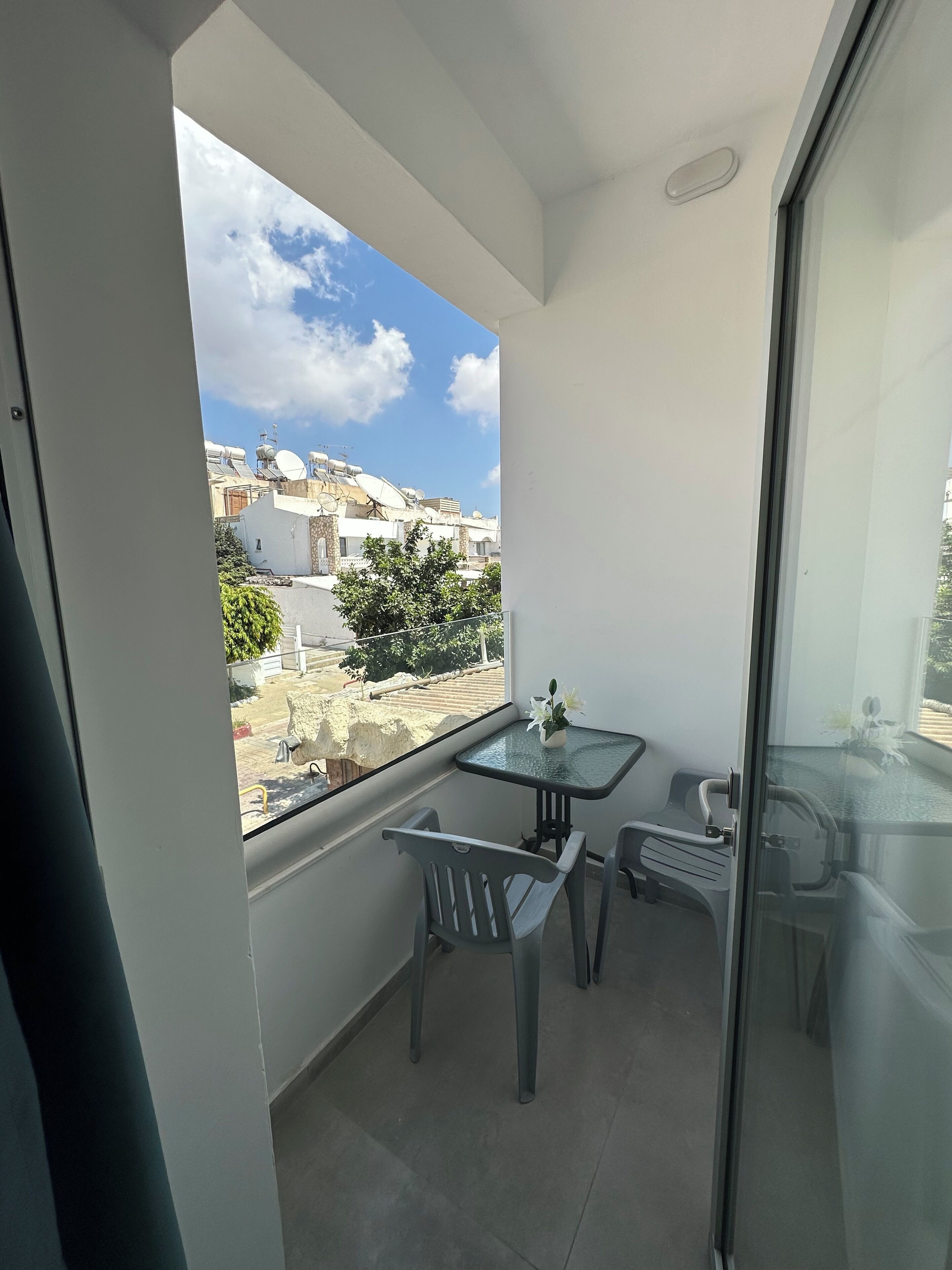 Un balcon avec une table en verre et deux chaises, une petite plante en pot, et une vue sur les bâtiments et les arbres voisins.