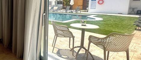A view from a hotel room to an outdoor pool area with chairs and a table, a grassy lawn, and a building in the background.