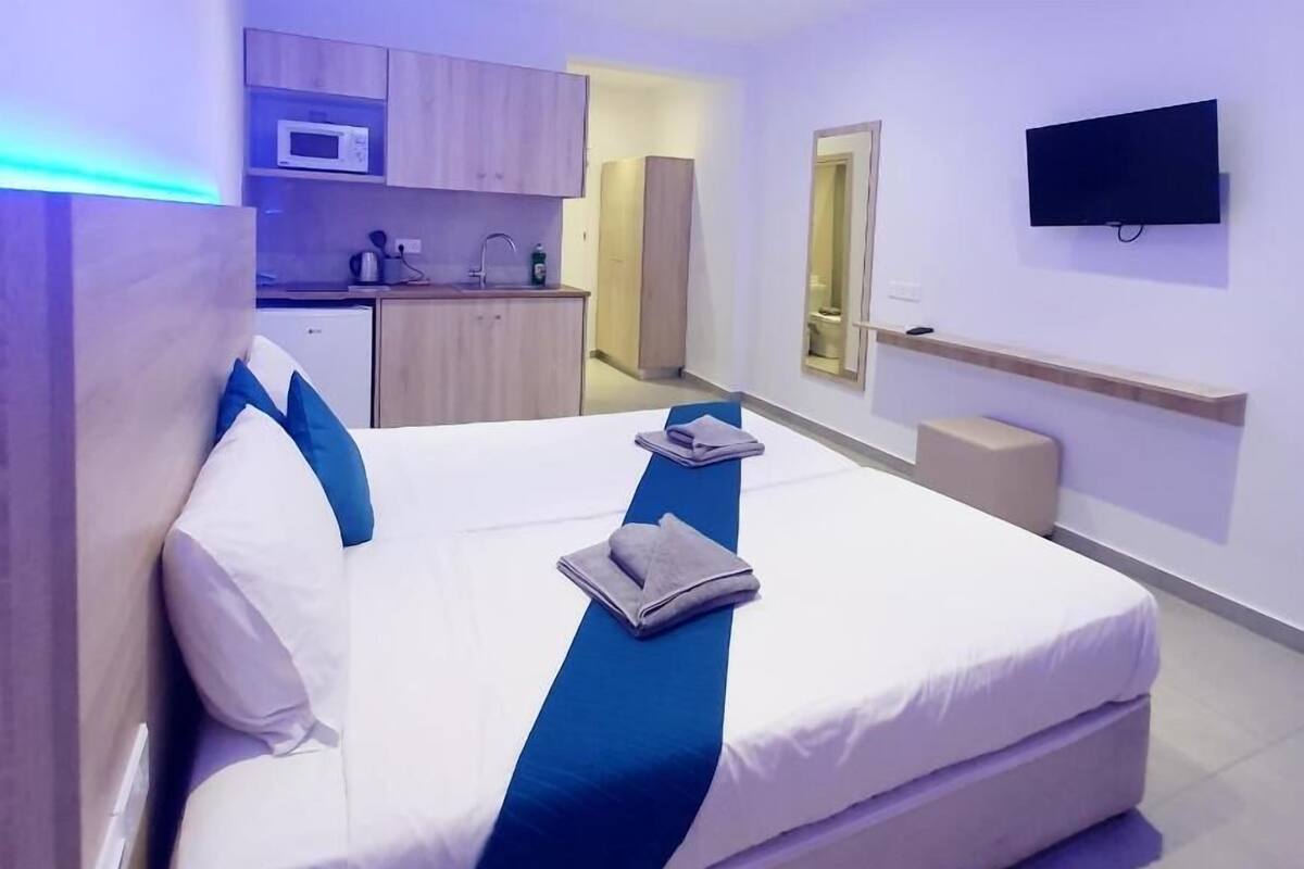 A modern hotel room with a large bed, a flat-screen TV mounted on the wall, and a kitchenette area in the background.