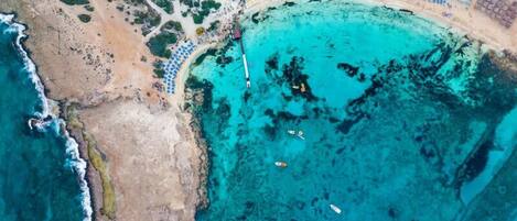 An aerial view of a coastal area with a beach, clear turquoise waters, and a series of blue umbrellas arranged in rows along the shore.