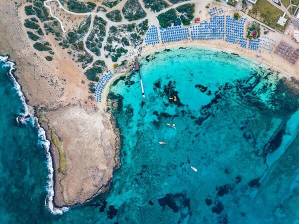 Vista aérea de una zona costera con una playa, aguas turquesas cristalinas y una serie de sombrillas azules dispuestas en filas a lo largo de la orilla.