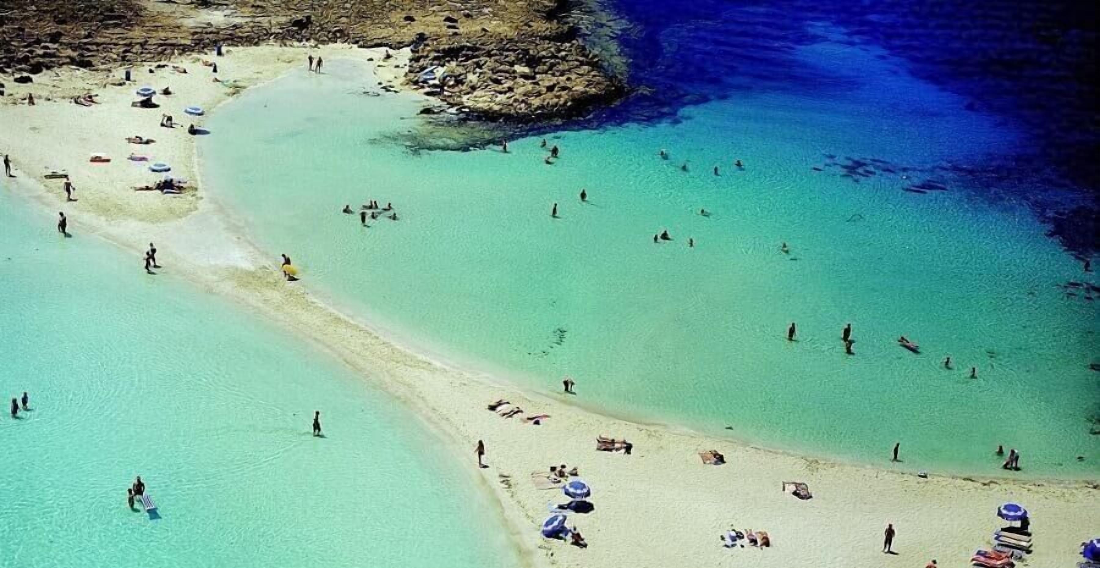 An aerial view of a beach with clear turquoise water, sandy shore, and people swimming and sunbathing.