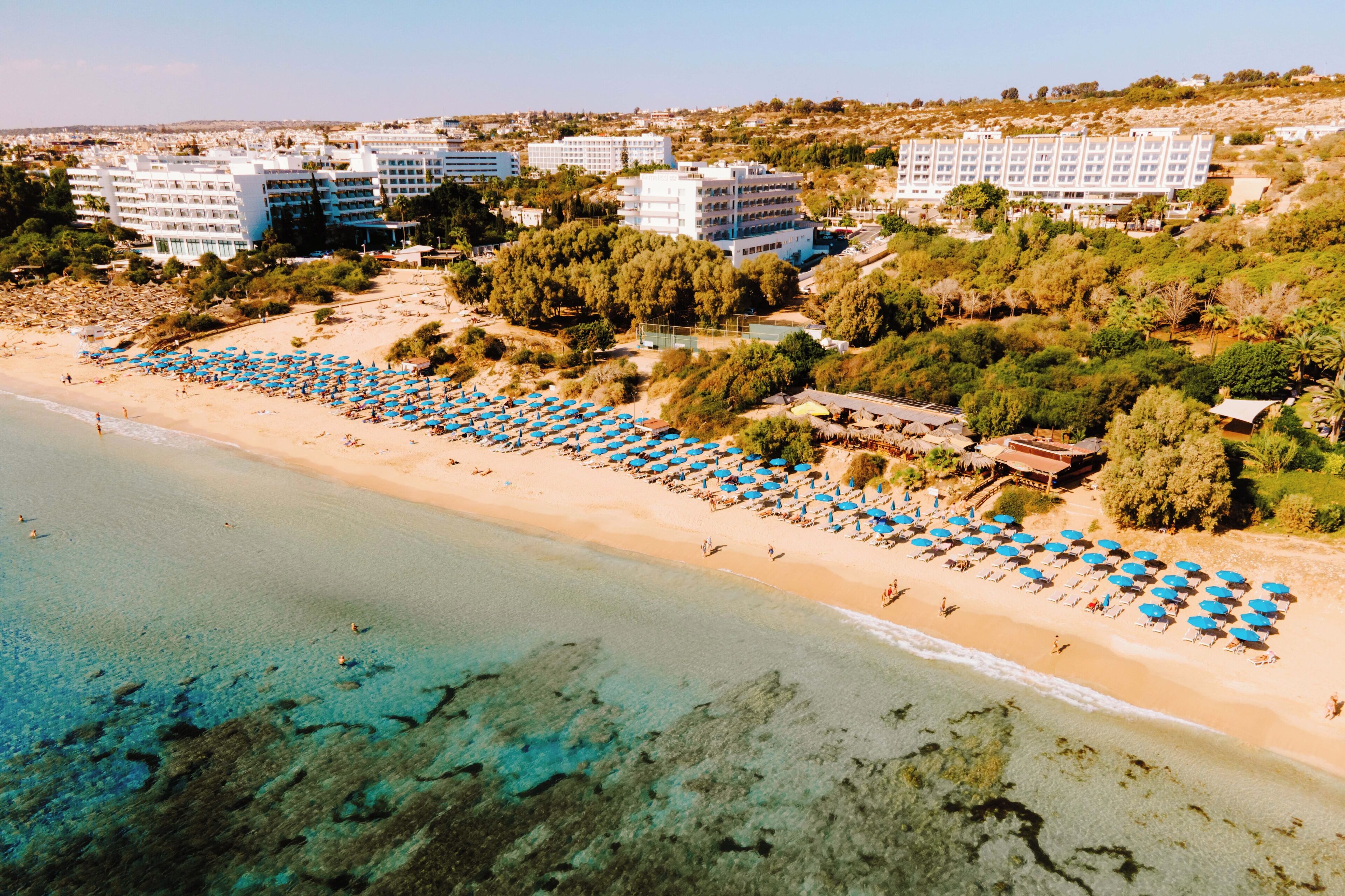 Un resort frente al mar con varios edificios de hotel, una playa de arena bordeada por sombrillas azules y aguas turquesas cristalinas.