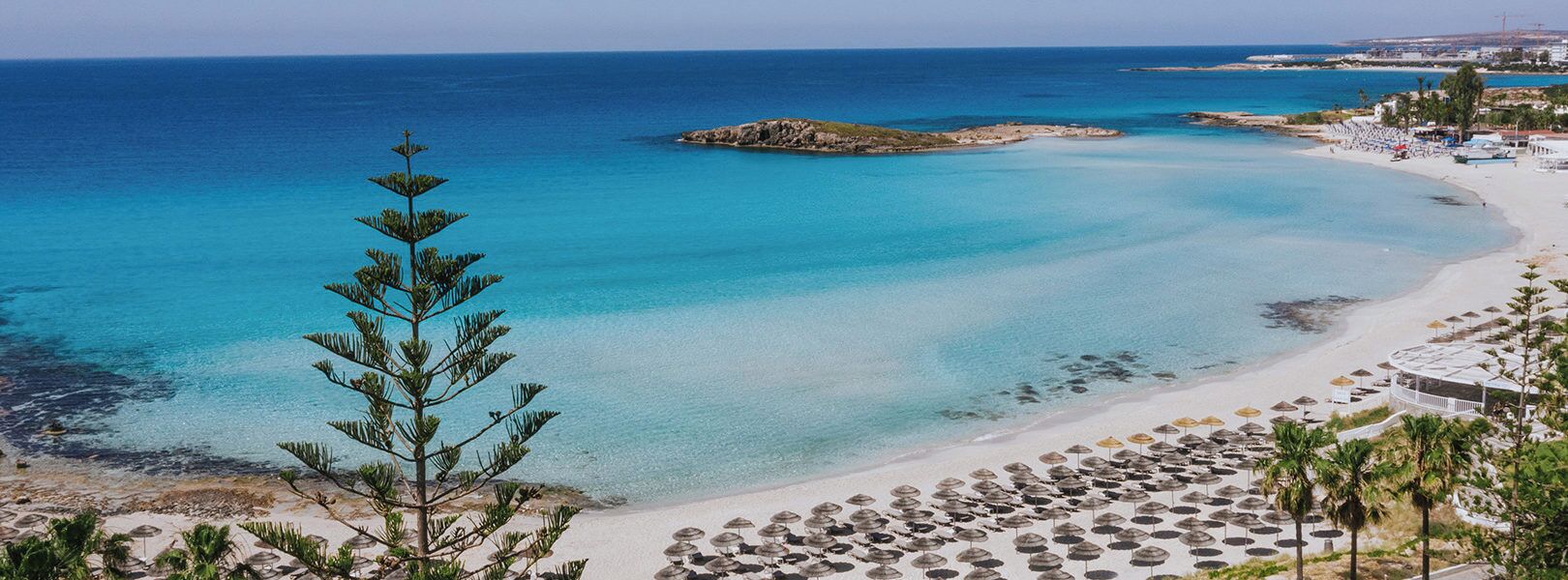 Un complejo turístico en la playa con filas de tumbonas y sombrillas, un mar azul cristalino y un promontorio rocoso en la distancia.
