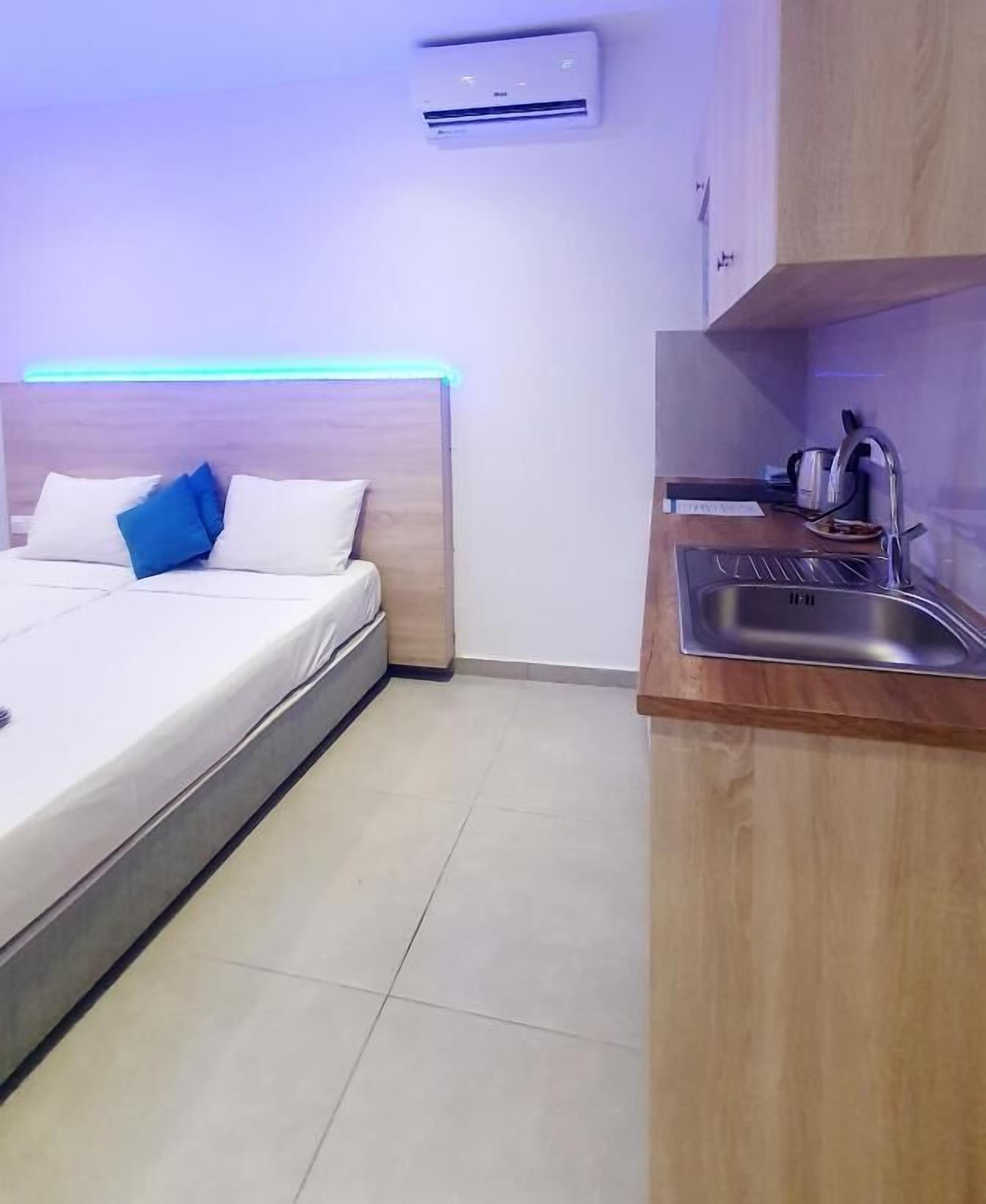 A small, modern room with a bed, a bedside table, a built-in sink, and an air conditioning unit.
