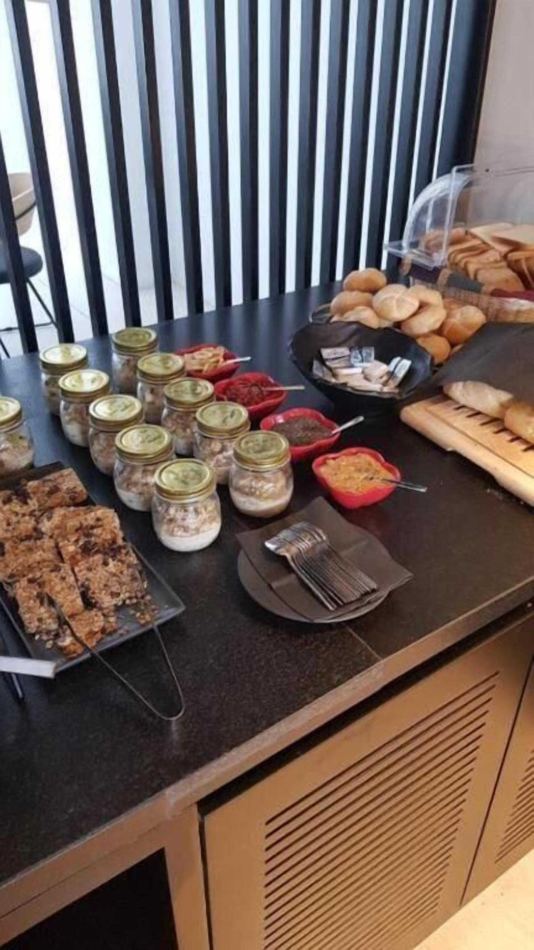Mesa de buffet com diversos alimentos, incluindo potes de condimentos, pão e uma bandeja de barras de granola.