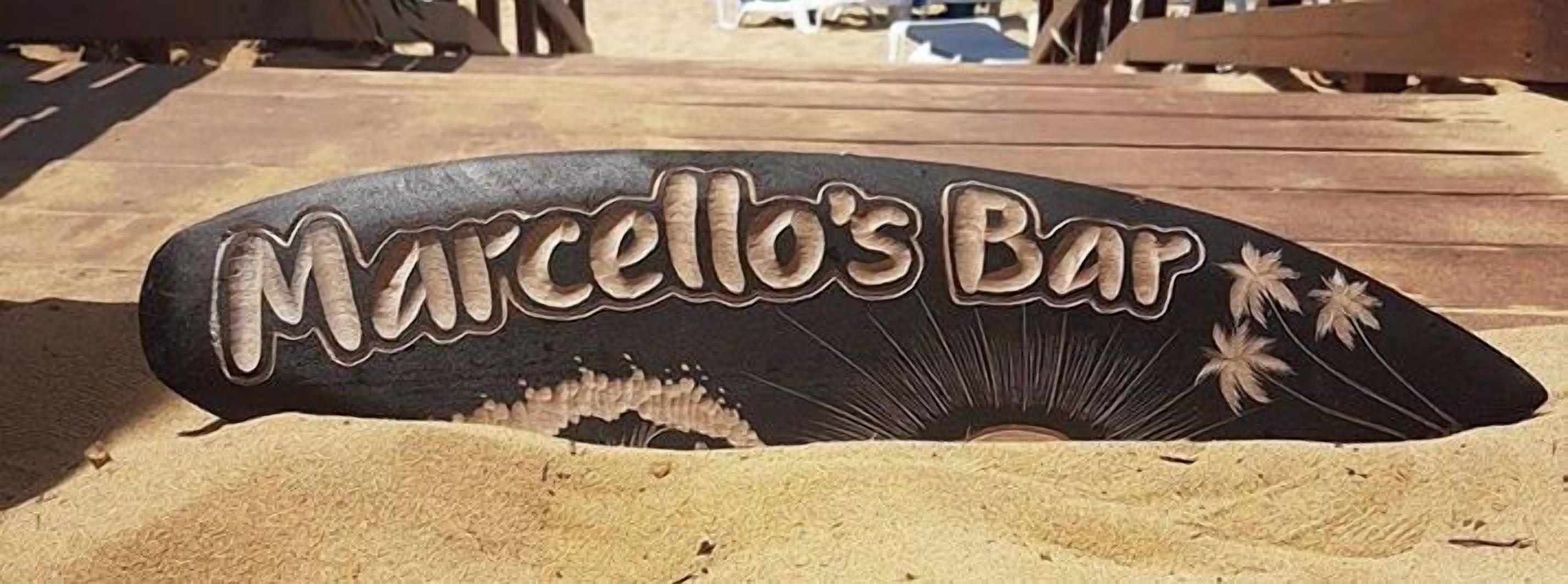 Een houten bord met de tekst 'Marcello's Bar' op een zandige ondergrond.