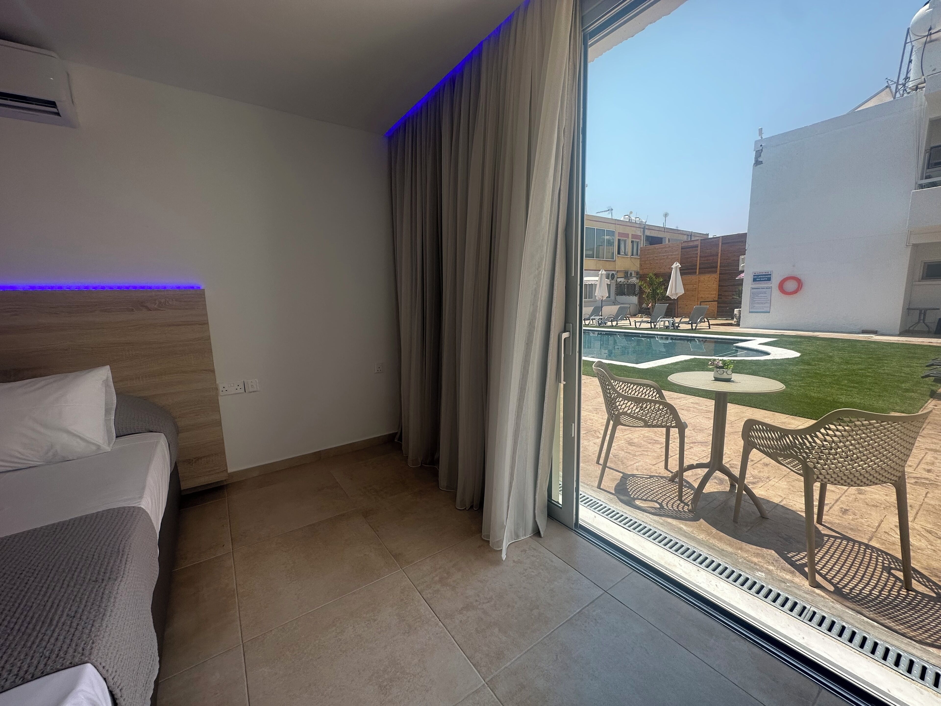 Camera d'albergo con un letto, una finestra con tende e vista su una zona piscina all'aperto con sedie e ombrelloni.