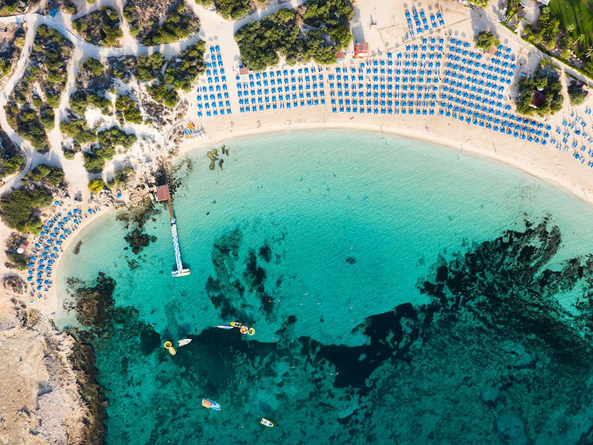 Vista aérea de una playa con hileras de sombrillas azules y un muelle que se adentra en las aguas turquesas.
