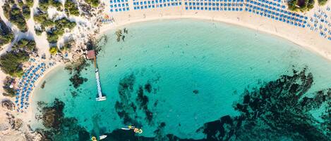 Vista aérea de una playa con hileras de sombrillas azules y un muelle que se adentra en las aguas turquesas.