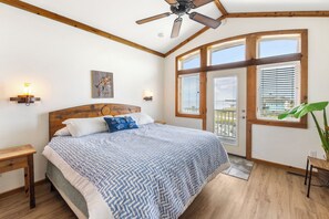 A bedroom with a bed, wooden headboard, two bedside tables, a ceiling fan, a window with blinds, and a view of the outdoors.
