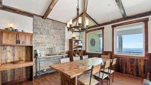Un coin repas rustique avec une table et des chaises en bois, un mur en pierre et une grande fenêtre donnant sur un paysage.