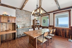 A rustic dining area with a wooden table and chairs, a stone wall, and a large window offering a view.